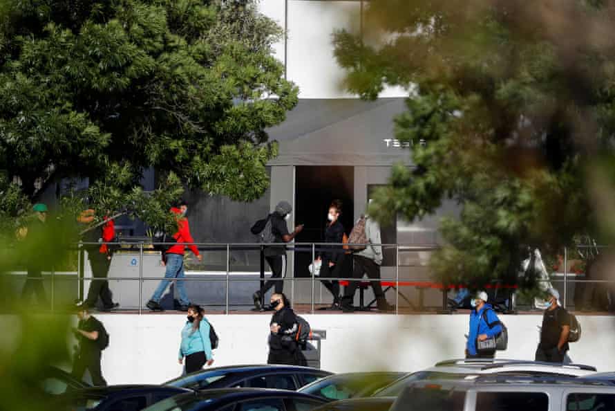 Workers wearing masks line up for a shuttle in front of a dark gray tent with "Tesla" on it.