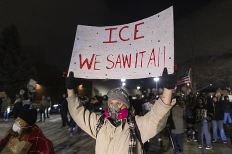 A protester wearing a beige jacket, gas mask, gray beanie and black gloves holds up a white sign that reads “ICE WE SAW IT ALL.” Behind the protester is a group of other protesters holding an American flag against a dark sky.