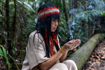 Adana Kambeba sitting on a log in the forest wearing a feather headdress and white robe