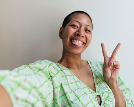 A selfie shot of a young woman wearing a checked summer dress, standing next to a window in a building in Newcastle upon Tyne. She is looking at the camera and sticking her tongue out while posing with the peace sign.
