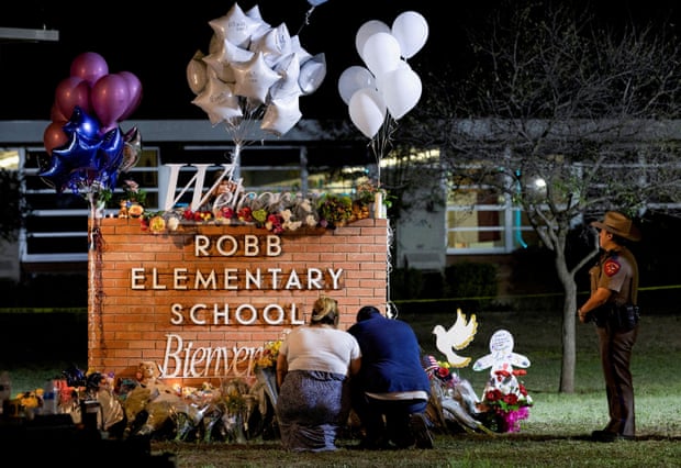 Two people kneel in front of a sign reading 'Robb elementary school' which is surrounded by bouquets of flowers and bunches of balloons. An officer stands off to the side.