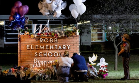 Two people kneel in front of a sign reading 'Robb elementary school' which is surrounded by bouquets of flowers and bunches of balloons. An officer stands off to the side. Two people kneel in front of a sign reading 'Robb elementary school' which is surrounded by bouquets of flowers and bunches of balloons. An officer stands off to the side.