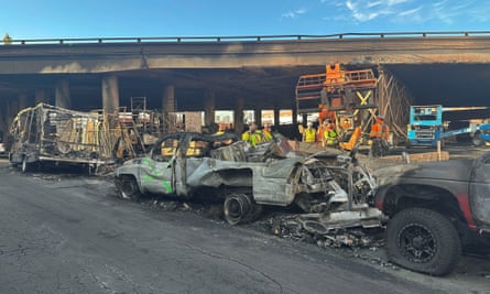 Workers in yellow and orange safety vests among burned-out vehicles below an overpass.