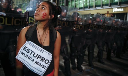 A protester standing before a police barricade, her face painted with the female gender symbol, bears a sign reading: ‘Rape never more’