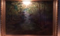 A picture of a framed painting. There is a glare from the flash of a camera at the top in the middle of the painting. It depicts a stream running through a forest.