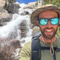 A man wearing reflective sunglasses and a sunhat stands in front of a waterfall coming down over granite, on a sunny day.