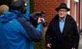 George Galloway gives an interview to a TV station in Wardleworth, Rochdale: he wears a black hat, black overcoat and blue shirt, and is facing a cameraman who wears a blue anorak and is seen from behind. He stands in front of a terrace of red-brick houses; it looks as if the weather is wet and wintry.