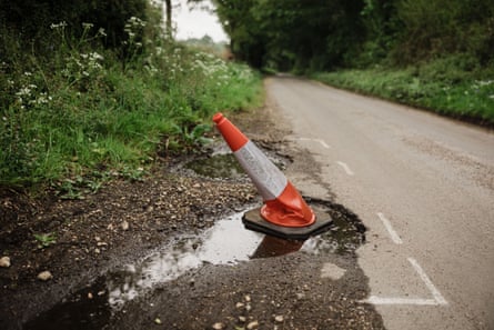 An orange and white traffic cone standing in a pothole at the side of a road