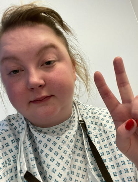 A woman with blond hair wearing a hospital gown, with her fingers making the ‘peace’ sign