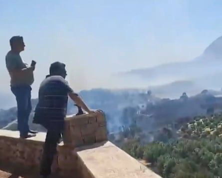 Screenshot of two people on balcony look out at parched landscape with smoke rising