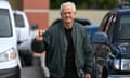 Older white man with white hair, wearing black T-shirt and olive bomber jacket, smiles and gives peace sign as he walks through parking lot among cars.