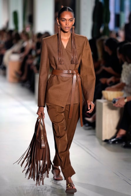 A model wearing a brown trouser outfit with a belted jacket, long leather earrings and a handbag covered in fringes