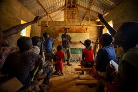 A teacher stands in front of a class of children in a small building