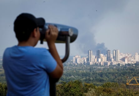 A boy looks through binoculars at Kyiv in the distance, where smoke rises over city buildings.