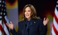 woman wearing dark blue suit gestures behind microphone with two US flags behind her