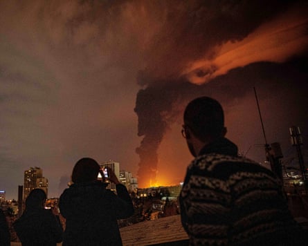 Three silhouetted people watch a dark smoke plume rise from a fire on the horizon of Tehran