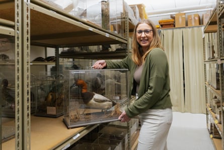 A smiling woman holding plexiglass box with a bird in it alongside shelving units.