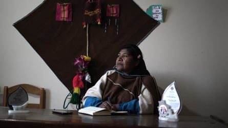 An Indigenous Latin American woman sitting at a desk with woven goods attached to the wall behind her