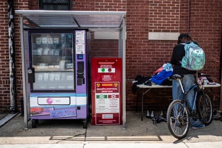 A person with a backpack and a bicycle stands at a table piled with items, next to a large vending machine and what looks like a clean needle dropoff box.