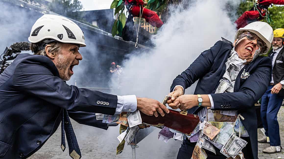 Climate activists in suits and hard hats wrestle over a briefcase containing banknotes