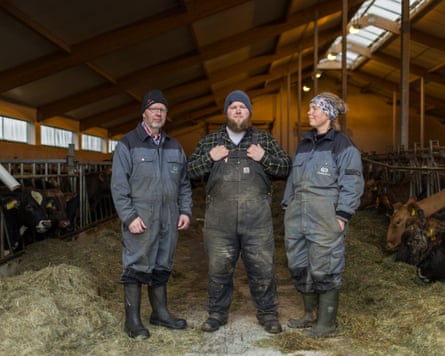 The three farmers, all wearing heavy-duty overalls and wellies, stand among their dairy herd in the cowshed, with young bulls visible. The two men, who both have beards, wear woolly hats and Davíð’s mother has a scarf tying back her blond hair.