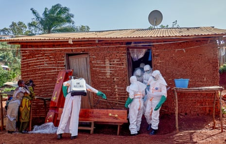 A person in PPE prepares a coffin while four others carry a body from a small mud house. Two grieving women stand to the left of them.