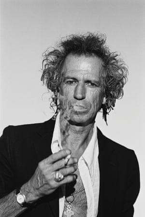 Keith Richards in a white open shirt and black jacket, holding a cigarette and smoke coming from his mouth