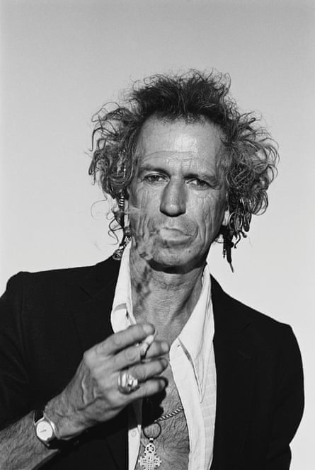 Keith Richards in a white open shirt and black jacket, holding a cigarette and smoke coming from his mouth