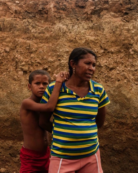 A woman in a striped shirt stands with a young boy, who is holding on to her shoulder with both hands.