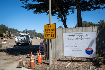 Large sign on side of wooden fence indicating building project, alongside orange cones and white truck.