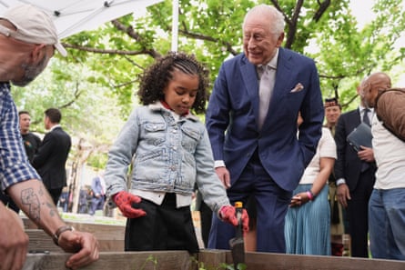 A man in a blue suit, standing next to a young girl wearing gloves and digging in a dirt planter.