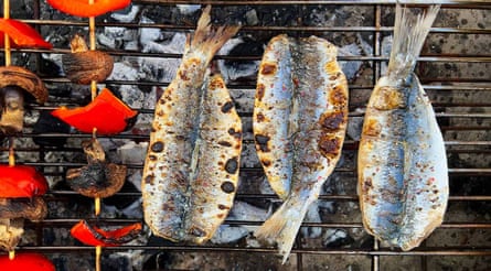 Three fish cooking on a barbecue alongside some red peppers and mushrooms on skewers.