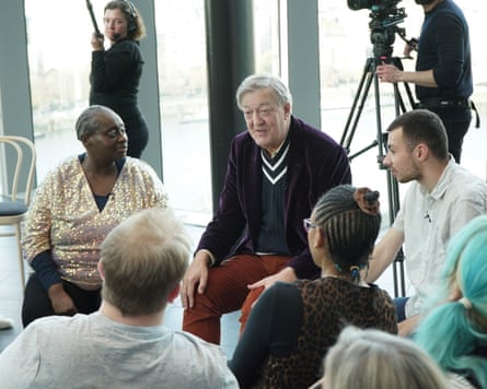 A seated grey-haired man in purple velvet jacket, blue and white stripe jumper and red cord trousers with hands on knees talking to a group of young people around him.