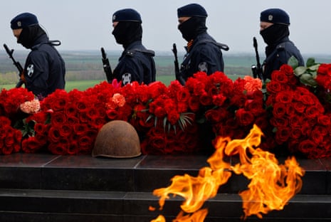 Four uniformed members of the Russian National Guard face left as they stand behind a row of flowers and a burning flame. The members all have their faces covered and are holding weapons.