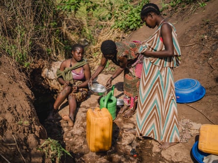 Three women fill water cans from a muddy stream