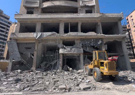 A yellow bulldozer sits in front of the debris and wreckage of a tall building set against a blue sky.
