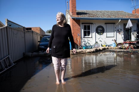 Carol Adams is standing in her driveway with flood water around her bare feet. The flood waters have just reached the sandbags along the front of her house behind her