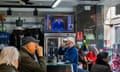 Customers sit at tables at a cafe, some of them drinking coffee, as a TV mounted above a fridge shows Pedro Sánchez delivering an address