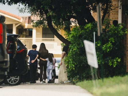 A group of people, including children, seen from behind, walking down a tree-lined sidewalk with their arms wrapped around each other.