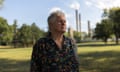 An older person looks wistful and frustrated as she stands on a grassy stretch of land with trees, while fumes rise out of a power plant in the distance