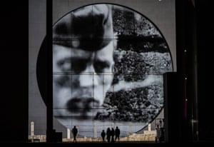 The soldier's face appears in closeup in black and white within the circular projection, as a few people stand below watching the film
