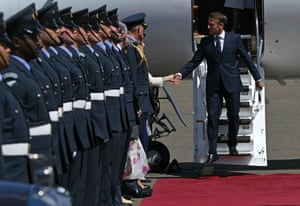 A man a suits walks down the steps of a plane on onto a red carpet lined with military personnel in ceremonial unforms