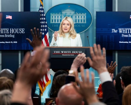 Karoline Leavitt speaks at a lectern while journalists in the foreground raise their hands