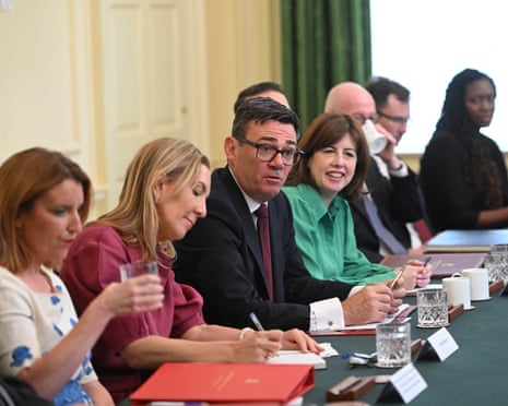 Prime minister Andy Burnham sits at a cabinet meeting table with colleagues, taking notes.
