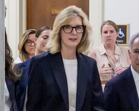 A middle-aged woman in glasses walks in a corridor surrounded by other people.