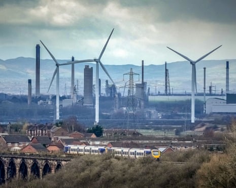 wind turbines set amid an industrial plant as a train runs through the foreground