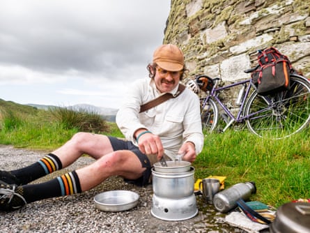 A man in shorts, cap and natty socks stirs food in a camping stove, with his bike resting on a stone wall behind