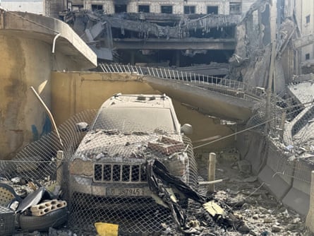 A vehicle in the foreground is covered in dust and rubble, with a destroyed building behind.