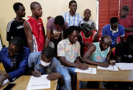 A group of men fill in forms with other men standing behind them.