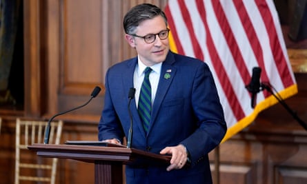 A man wearing a blue suit and glasses is standing behind a lectern and looking toward his side